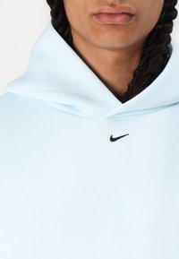Ανοιχτό μπλε φούτερ με μαύρο λογότυπο Nike στο στήθος. Διαθέτει κουκούλα με κορδόνι και λεία υφή. Χωρίς ορατά σχέδια ή λεπτομέρειες.