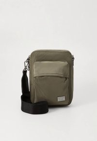 Malá olivově zelená kabelka přes rameno s předním klopovým kapsou, černým nastavitelným popruhem a pravoúhlou kovovou logovou destičkou v pravém spodním rohu.