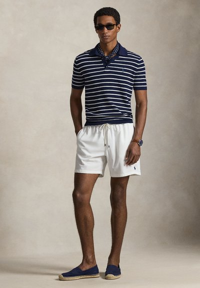 Πολύ μπλε και λευκό πουκάμισο πόλο με κοντά μανίκια, συνδυασμένο με λευκό σορτς και ναυτικά εσπαντρίγιες. Αξεσουάρ περιλαμβάνουν γυαλιά ηλίου και ρολόι.