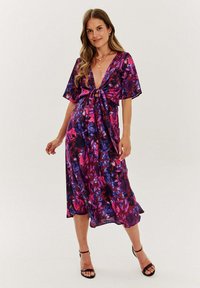 Kwiecista sukienka midi z głębokim dekoltem w kształcie V, krótkimi szerokimi rękawami oraz wiązaniem w talii. Materiał jest przezroczysty w odcieniach purpurowym i różowym, z ciemnymi kwiatami jako akcentami.