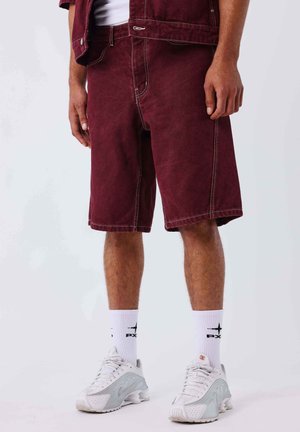 Mann trägt bordeauxrote knielange Shorts, weiße Socken mit Aufdruck und weiße Sportschuhe, steht vor einem schlichten hellen Hintergrund.
