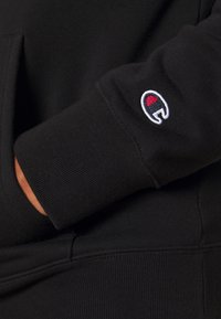 Manche de sweat-shirt noire avec poignet côtelé présentant un petit logo Champion brodé en rouge, blanc et bleu marine près du poignet.