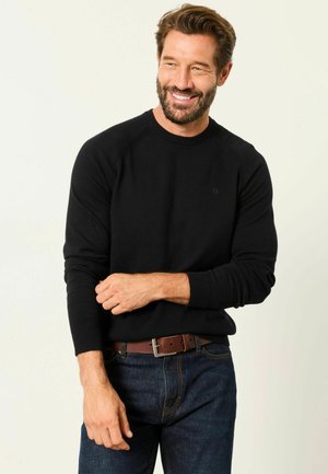 Mann mit Bart lächelnd, trägt schwarzen Pullover, blaue Jeans und braunen Gürtel, richtet den Ärmel vor schlichtem hellem Hintergrund.