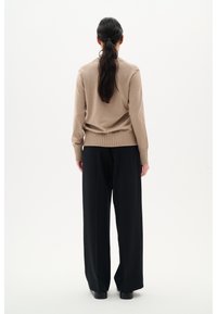 Maglione beige lavorato a maglia con orlo a coste abbinato a pantaloni neri a manica larga a vita alta, visto da dietro su uno sfondo bianco.