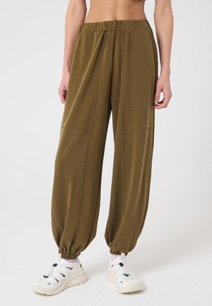 Persona che indossa pantaloni da jogging larghi verde oliva con polsini elasticizzati e scarpe da ginnastica bianche, in piedi contro uno sfondo semplice.