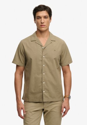 Giovane uomo con capelli corti e scuri indossa una camicia beige a maniche corte con bottoni e pantaloni abbinati, in piedi davanti a uno sfondo bianco.