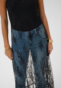 Schwarzes Spitzenoberteil mit floralen Mustern, über einem Jeansrock getragen. Ein goldenes Armband am Handgelenk setzt metallische Akzente.