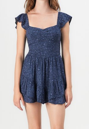 Kobieta ubrana w granatowy kombinezon w jasnoniebieskie kwiatowe wzory, z falbankowymi rękawkami i kwadratowym dekoltem, stojąca na jednolitym tle.