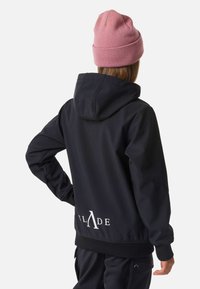 Giacca nera con cappuccio e tessuto morbido, con logo bianco "BLADE" sul retro e polsini a costine. Abbinata a un berretto rosa lavorato a maglia.