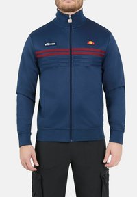 Veste zippée bleu marine en tissu lisse, dotée de rayures rouges horizontales, d'un col montant et d'un logo sur la poitrine.