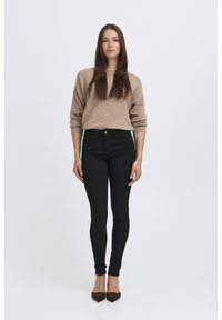 Pull en tricot marron clair associé à un jean skinny noir taille haute et des escarpins noirs à bouts pointus, présenté sur un fond blanc.