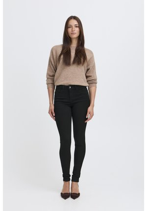 Hellbrauner gestrickter Pullover kombiniert mit hochtaillierten schwarzen Skinny-Jeans und dunklen spitzen Absatzschuhen, vor einem weißen Hintergrund präsentiert.