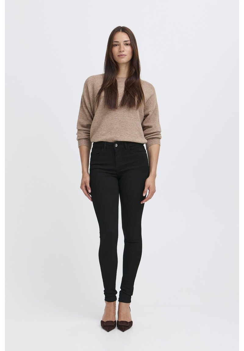 Pull en tricot marron clair associé à un jean skinny noir taille haute et des escarpins noirs à bouts pointus, présenté sur un fond blanc.