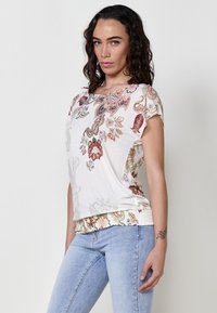 Haut blanc à manches courtes avec un motif paisley multicolore, ourlet élastique et texture douce, associé à un jean ajusté bleu clair.
