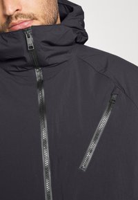 Svart huoodad jacka med strukturerad tyg, som har två diagonala fickor med dragkedja och en hel dragkedja fram.