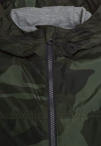 Veste à motif camouflage, présentant une fermeture à glissière intégrale, une doublure intérieure grise, et un tissu léger et brillant avec une texture lisse.