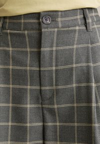 Pantaloni a quadri grigi realizzati in un tessuto testurizzato, con un bottone visibile e linee beige a contrasto in un motivo a scacchi.