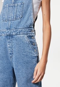 Jeanslatzhosen in Hellblau mit einer strukturierten Oberfläche, ausgestattet mit einer Vordertasche, seitlichen Knöpfen und einem lockeren Schnitt.