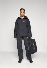 Zwarte waterdichte jas met rode accenten, gecombineerd met bijpassende broek met een patroon. De persoon houdt een zwarte rugzak vast.