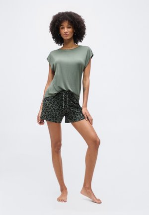 Femme debout pieds nus portant un t-shirt vert fluide et un short à imprimé léopard vert foncé sur un fond uni.