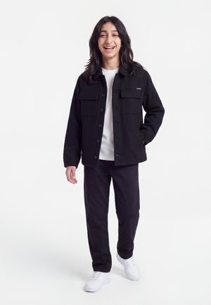 Young person smiling, wearing a black button-up jacket, white shirt, black pants, and white sneakers, standing against a plain white background.