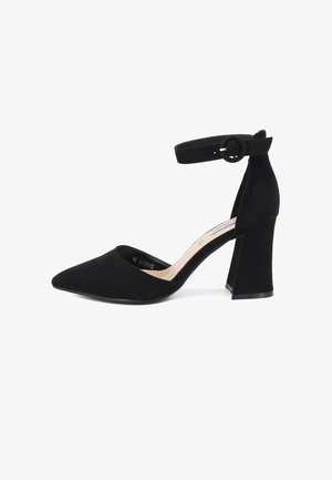 Tacchi alti in suede nero con punta affusolata e cinturino alla caviglia dotato di una fibbia circolare. Il tacco è spesso e strutturato, migliorando la stabilità.