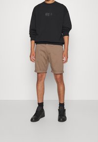 Felpa nera con logo impresso, abbinata a pantaloni corti marroni a risvolto e sneakers nere. Design semplice, materiale in cotone e vestibilità casual.