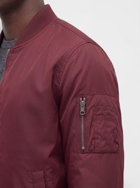 Homme portant une veste bomber bordeaux avec une poche zippée sur la manche supérieure et un col côtelé.