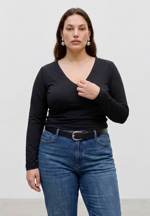 Femme aux longs cheveux bruns portant un haut noir à col en V à manches longues, un jean bleu avec une ceinture noire et des boucles d'oreilles argentées remarquables, debout devant un fond uni.