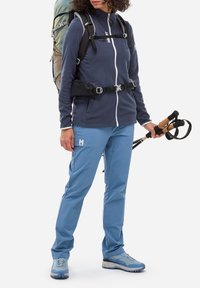 Tenue de plein air comprenant une veste polaire bleu foncé avec une fermeture éclair blanche, un pantalon imperméable bleu clair et des chaussures de randonnée bleues. Sac à dos ajustable et bâtons de randonnée.