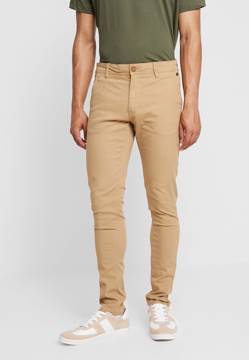 Homme portant un pantalon beige slim, des baskets blanches et beiges, et un t-shirt vert olive, debout devant un fond uni.