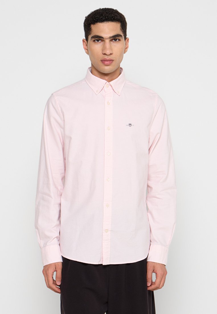 Ein junger Mann steht selbstbewusst da, trägt ein hellrosa Hemd mit Knöpfen und einem kleinen Logo auf der Brust vor einem schlichten weißen Hintergrund.