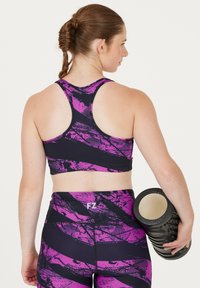 Svart sportbehå och leggings med ett lila abstrakt mönster. Personen håller en svart skumrulle i ena handen.