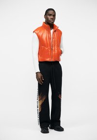 Άντρας που φοράει φωτεινό πορτοκαλί επενδεδυμένο γιλέκο πάνω από λευκό μακρυμάνικο πουκάμισο, μαύρο παντελόνι με εκτύπωση φλόγας και λευκό κείμενο στις πλευρές, μαύρα παπούτσια.