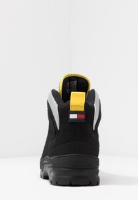 Černá semišová bota s gumovou podrážkou, šedými akcenty a žlutým poutkem. Na patě se nachází malý emblem v červené, bílé a modré barvě.