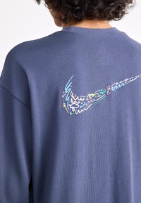 Sötétkék, laza fazonú pulóver, hátulján színes, absztrakt hímzett Nike logóval. Puha tapintás és laza dizájn jellemzi.