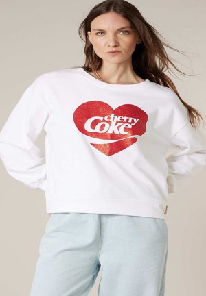 Femme aux longs cheveux bruns portant un sweat-shirt blanc avec un grand cœur rouge et le texte "cherry Coke", associée à un pantalon bleu clair.