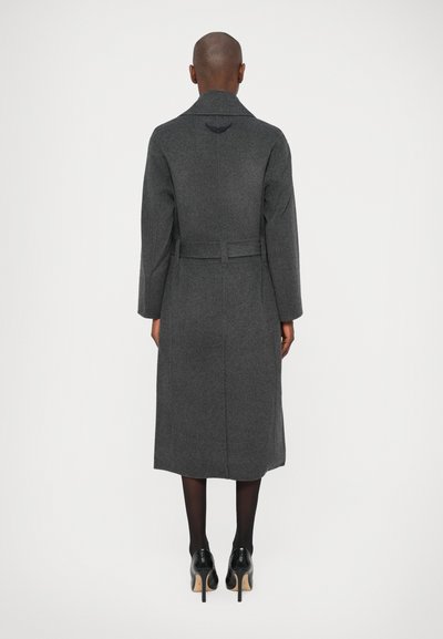 Szürke gyapjú trench coat övvel a deréknál, hátul szellőző résszel és finom hímzett részletekkel a gallér közelében. Fekete magassarkú cipők.