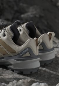 Scarpe da trail Adidas Terrex beige e grigie su una superficie rocciosa, che mostrano suole testurizzate e linguette sul tallone.