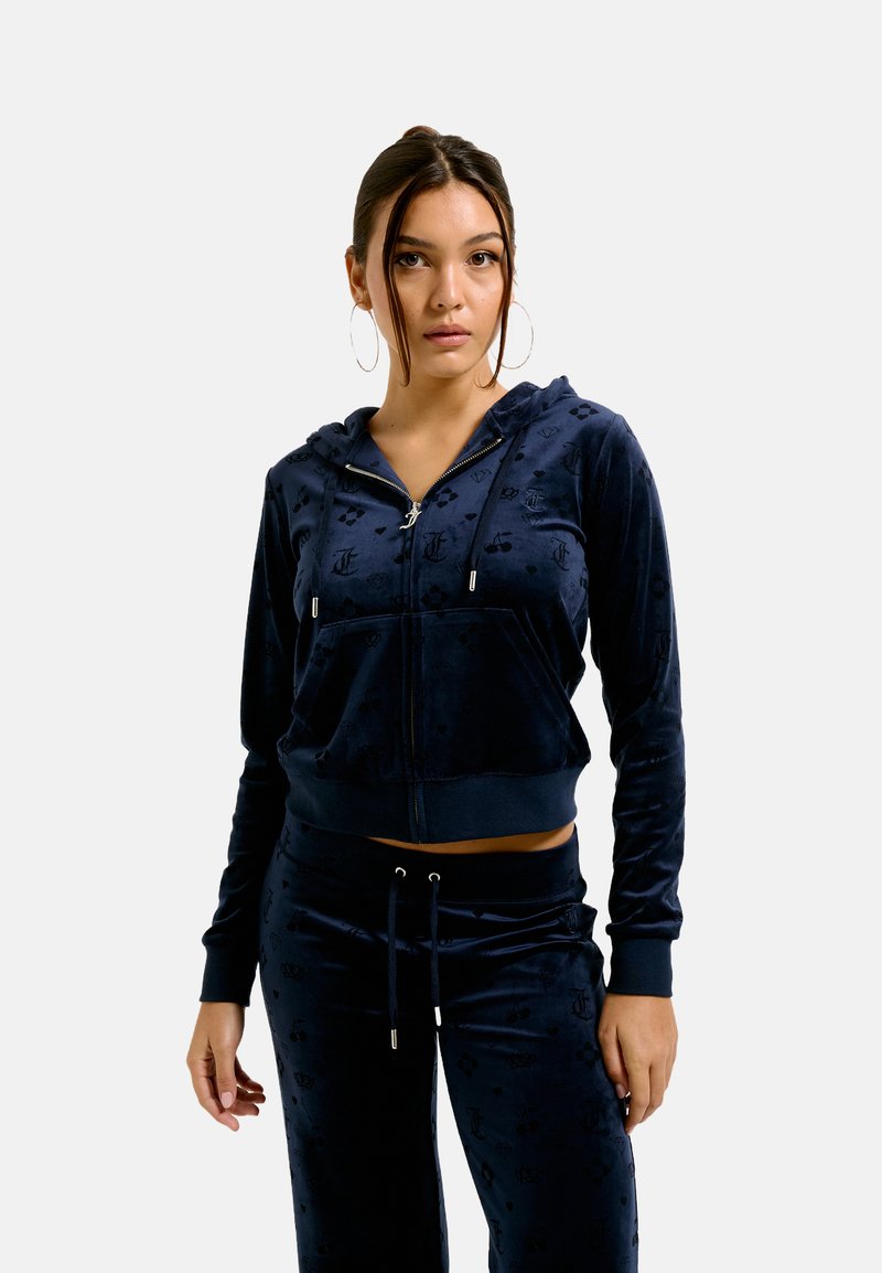Navy blue velour zip-up hoodie with logo pattern, front pockets, and drawstring hem. Paired with matching joggers, featuring similar design.