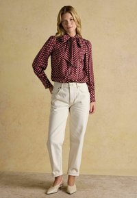 Burgundy polka dot blouse with a bow tie collar, paired with cream high-waisted jeans and light beige heeled mules on a textured background.