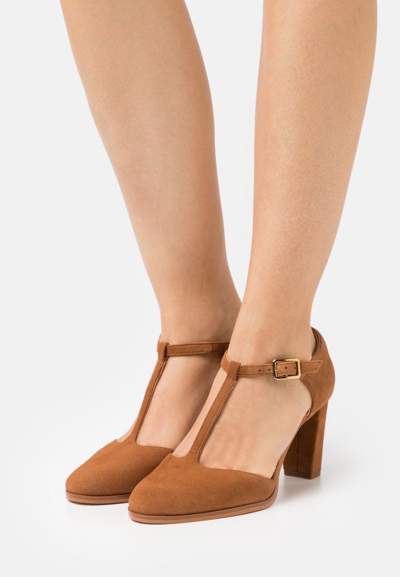 Tacchi alti in suede marrone con cinturino T e tacco a blocco, dotati di chiusura con fibbia e punta appuntita, su uno sfondo neutro.