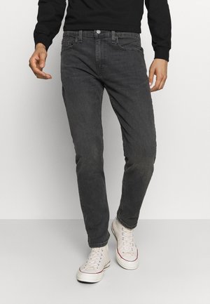 Homme portant un jean gris foncé, une chemise noire à manches longues et des baskets blanches montantes, debout devant un fond uni.