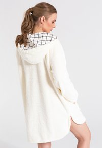 Biały, puszysty szlafrok z kapturem i podszewką w wzór kratę. Posiada długie rękawy i boczne rozcięcia, zapewniając luźny krój.