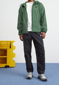 Green zip-up jacket with a hood, black logo, paired with black cargo pants and grey hiking shoes. Smooth fabric and practical design details.