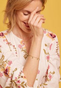 Mujer con cabello corto y rubio sonriendo y cubriendo parte de su rostro con la mano, vistiendo una blusa floral y una pulsera dorada con charms verdes.