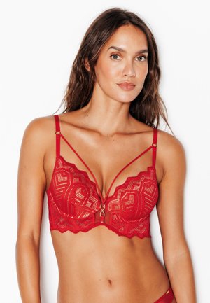 Donna con lunghi capelli castani che indossa un reggiseno in pizzo rosso con spalline decorative e motivi geometrici, posando davanti a uno sfondo bianco.