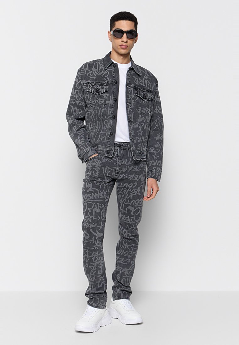 Hombre que lleva una chaqueta de mezclilla negra con estampado de graffiti y pantalones a juego, zapatillas blancas, camiseta blanca y gafas de sol negras, de pie sobre un fondo blanco.