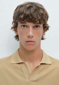 Polo shirt beige, testurizzato, con colletto e motivo a nido d'ape. Il modello ha capelli castani disordinati e tonalità di pelle neutre.