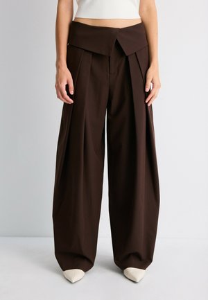 Pantalon large marron foncé avec une ceinture repliée, porté avec un top court blanc et des chaussures blanches pointues.